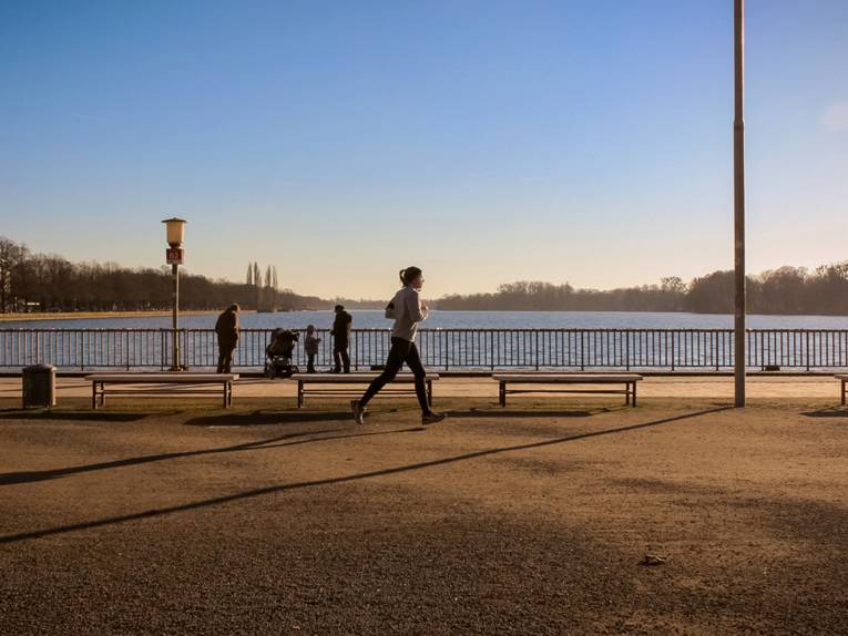 Eine Joggerin und drei weitere Personen vor dem Gitter des Maschsee-Nordufers.